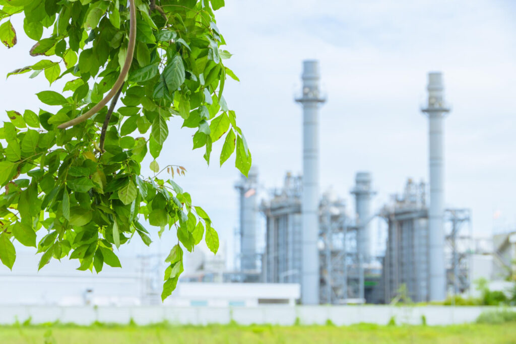 Green leaves in the foreground with an industrial plant in the background, symbolizing the environmental sustainability benefits of installing fixed gas detection systems.