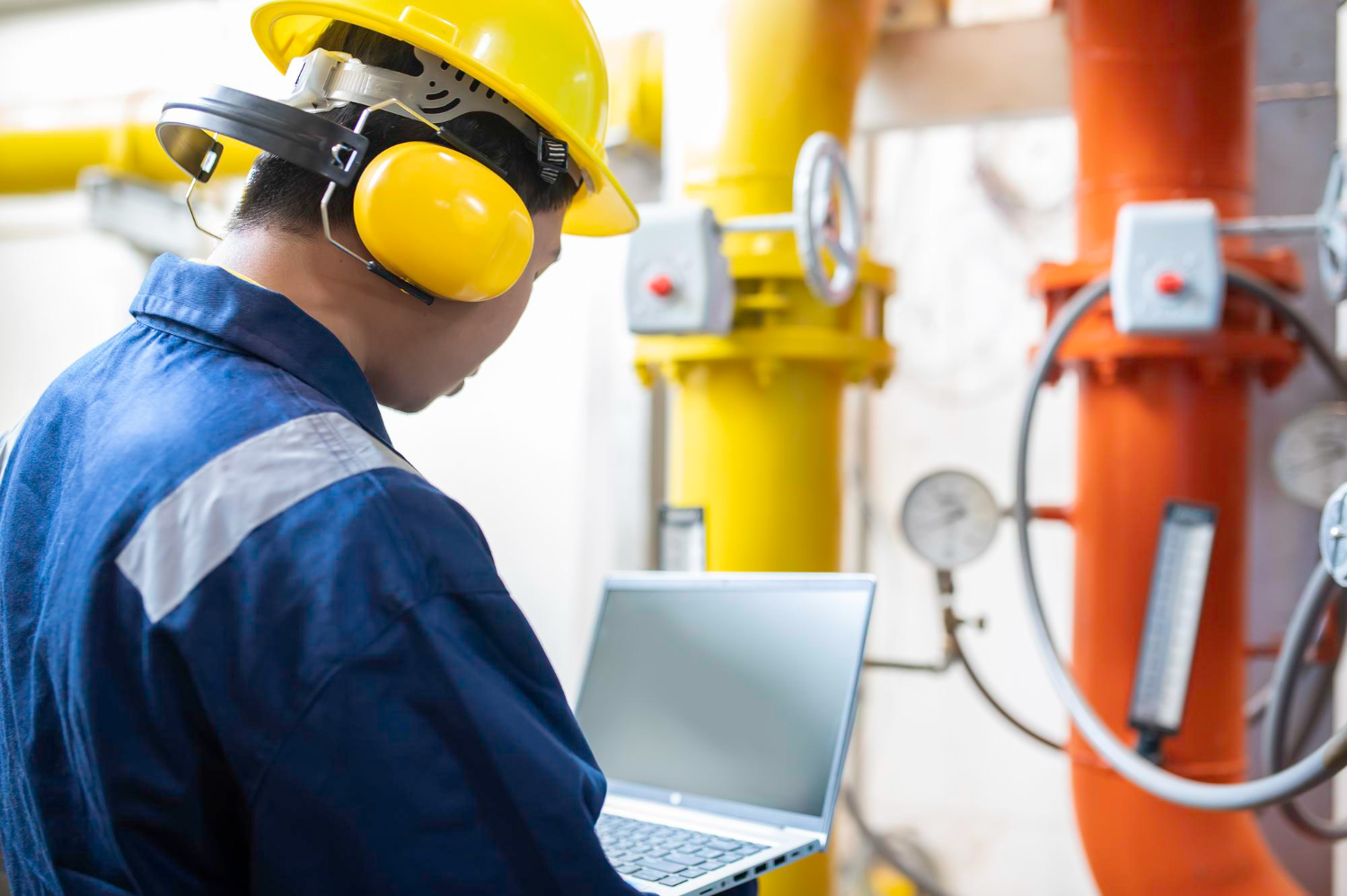 Technician using a laptop near industrial piping for methane gas detection and equipment safety monitoring in a facility.